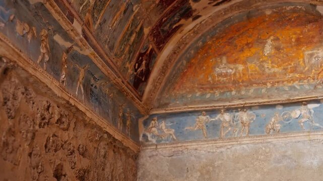 Upward view of a decorated Pompeii ceiling corner with blue fresco panels, painted human figures, and weathered Roman wall art preserved inside an ancient interior.