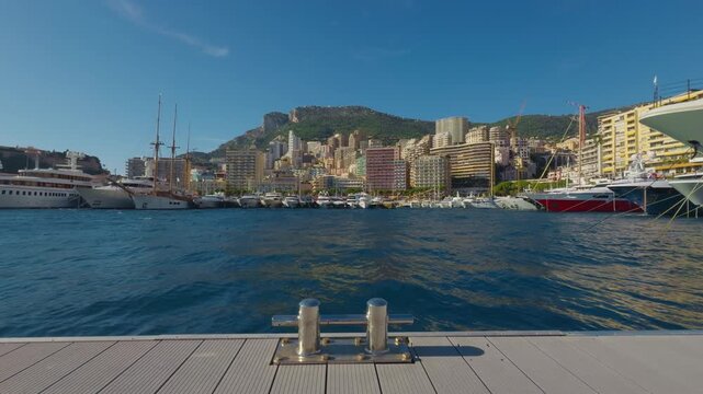 Static daytime shot from a pier in Port Hercule, Monaco. A metal mooring cleat in the foreground frames the luxury superyachts and dense La Condamine cityscape.