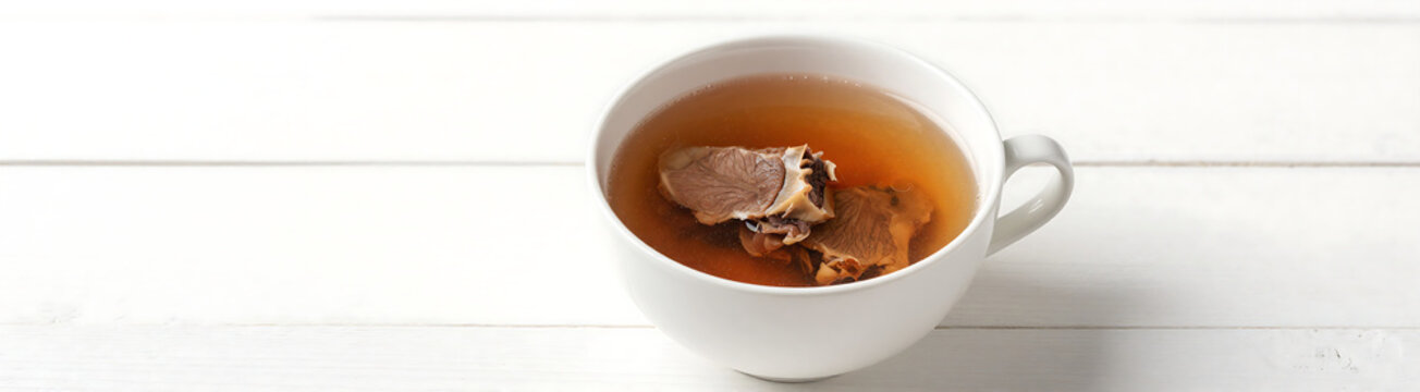 cup rich brown homemade beef bone broth sits atop clean white wooden surface surrounded empty space soft natural light