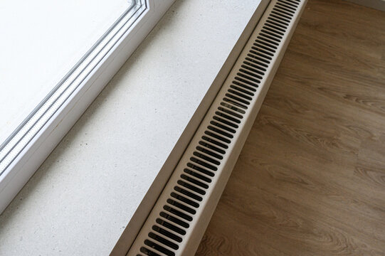 sleek floor-mounted convector heater placed under a large window, set against a light wall, with wooden flooring nearby; minimalist design, functional heating solution
