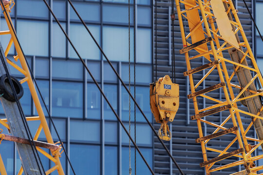 Yellow crane boom with 50-ton crane block and hook suspended by steel cables towers above urban development site adjacent to modern glass‑paneled building visible in background.