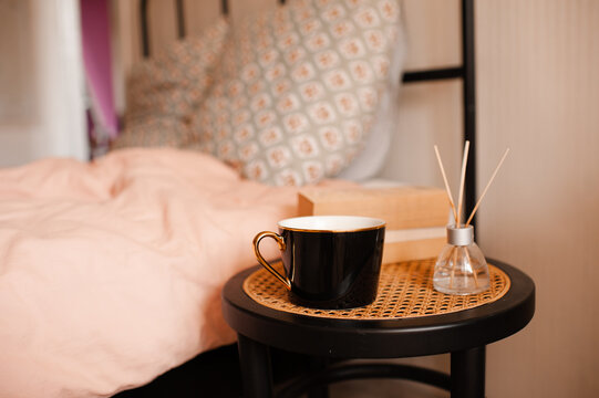 Cup of tea with aroma home fragrance in glass bottle with paper book stack on rattan table over bed in bedroom close up. Cozy domestic interior.