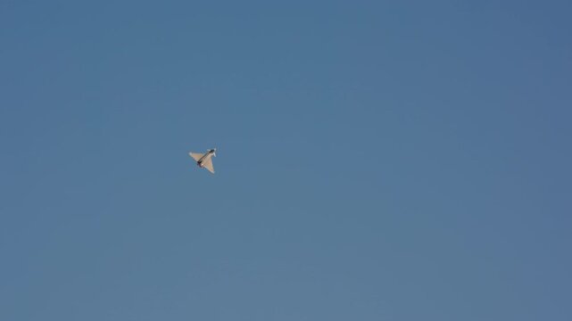 Military fighter jet executing steep climbing turn with visible afterburner glow against pale blue sky, slow motion, rear quarter view