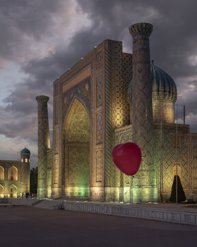 View of the Sher-Dor Madrasah with its intricate tilework and minarets under a dramatic twilight sky with a red heart-shaped balloon in Samarkand, Samarqand Region, Uzbekistan.