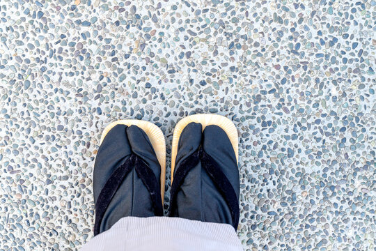 High angle view of a person wearing traditional Japanese black tabi socks and wooden sandals standing on a pebble stone floor. Authentic cultural footwear and urban outdoor texture background.