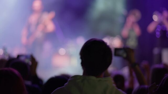 Unrecognizable fan from behind enjoys live music concert. Crowd dances and films with smartphones. Blurry stage with purple lights creates an energetic atmosphere. Shot from audience perspective