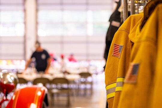 Bright yellow firefighter turnout coat hangs prominently at an indoor community event, figures and tables visible in the background in a scene of civic community life.