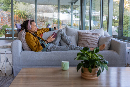 African American man reclining on sofa at home wearing mustard jacket, pink headset, holding phone