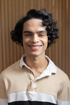 Teenage Indian male smiling wearing beige white shirt and hoop earrings in studio wooden-slat-wall