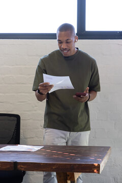 African American man standing behind wood table at office, in olive tee holding phone and paper