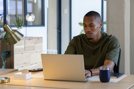 African American adult man wearing olive tee typing on silver laptop at office with smartphone