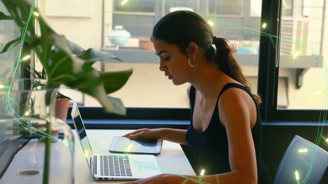 Woman sitting at counter by window, placing phone down then typing on laptop for remote work