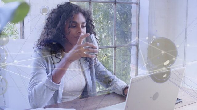 Woman typing on laptop, pausing, drinking and gazing out window with network overlaying for tech
