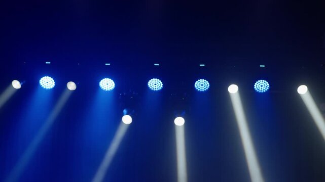 Wide row of bright blue LED par lights alternating with white moving head spotlights aimed downward, deep blue background fills upper frame