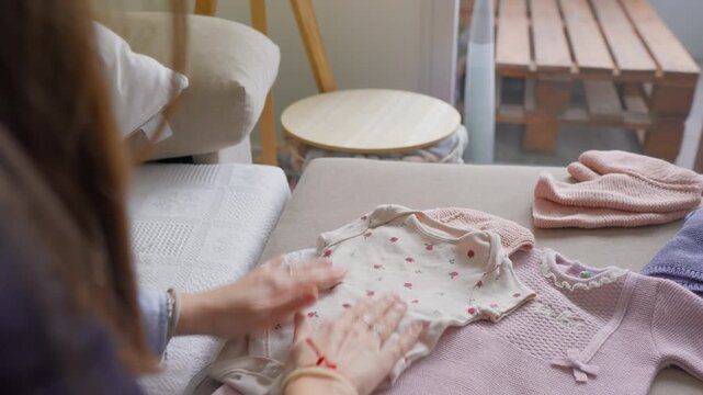 Expectant woman holds pink patterned baby bodysuit above bed surface with pink and mauve knitted garments nearby