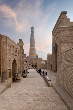 View of the Islam Khoja Minaret rising above narrow streets with people browsing market stalls and traditional brick architecture at sunset Khiva, Xorazm Region, Uzbekistan.