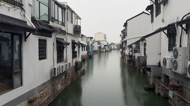 Serene canal waterway flowing between traditional Chinese whitewashed buildings in a historic water town, captured on a misty rainy day. Still water reflects the classic Jiangnan architecture with dar