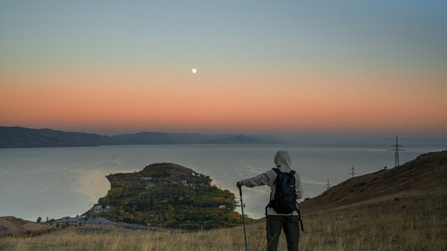 A lone hiker with a backpack stands on a hill overlooking Lake Sevan at sunset, gazing at the rising full moon above the tranquil water and distant mountains.