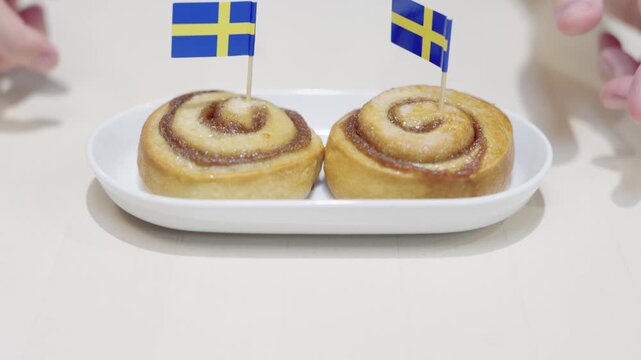 Traditional Swedish cinnamon rolls with national flags on a white plate. A person is reaching for a bun, showcasing Scandinavian pastry culture and fika tradition in a bright setting.