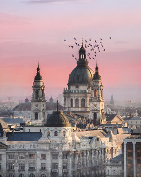 View of St. Stephen's Basilica with a flock of birds flying around the dome under a pink sunset sky in Budapest, Hungary.