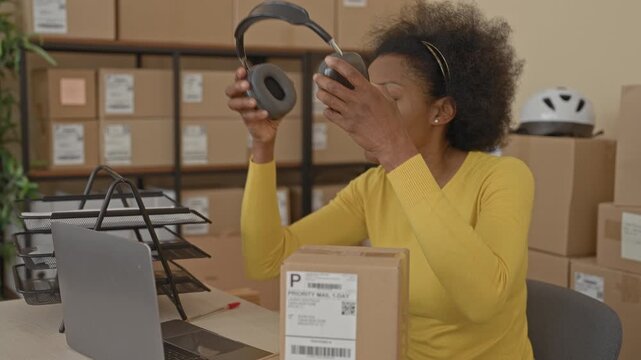Woman rubbing temple with hand near parcel and laptop amid stacked delivery boxes in a packing building with apparent headache; small business fatigue.