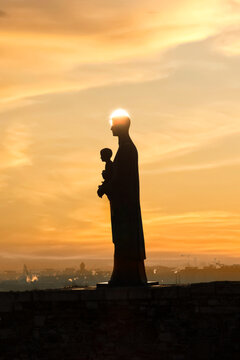 View of the silhouette of a religious statue holding a child with the sun creating a halo effect over a misty city skyline in Budapest, Hungary.