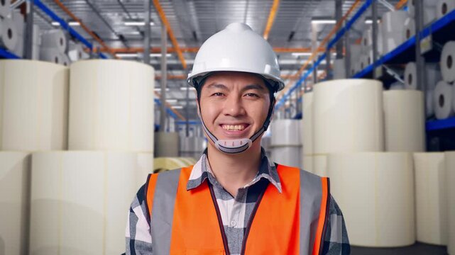Close Up Of Asian Male Engineer With Safety Helmet Smiling To Camera While Standing at Warehouse with Rows of Stacked White Polymer and Paper Rolls