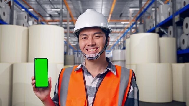 Close Up Of Asian Male Engineer With Safety Helmet Smiling And Showing Green Screen Smartphone To The Camera While Standing at Warehouse with Rows of Stacked White Polymer and Paper Rolls