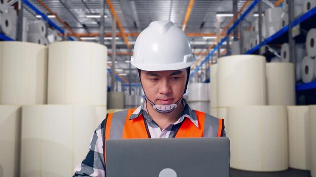 Close Up Of Asian Male Engineer With Safety Helmet Working On A Laptop While Standing at Warehouse with Rows of Stacked White Polymer and Paper Rolls