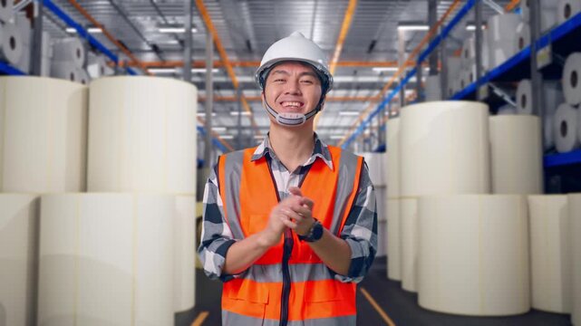 Asian Male Engineer With Safety Helmet Smiling And Clapping His Hands While Standing at Warehouse with Rows of Stacked White Polymer and Paper Rolls