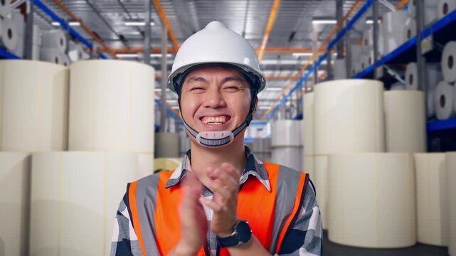 Close Up Of Asian Male Engineer With Safety Helmet Smiling And Clapping His Hands While Standing at Warehouse with Rows of Stacked White Polymer and Paper Rolls