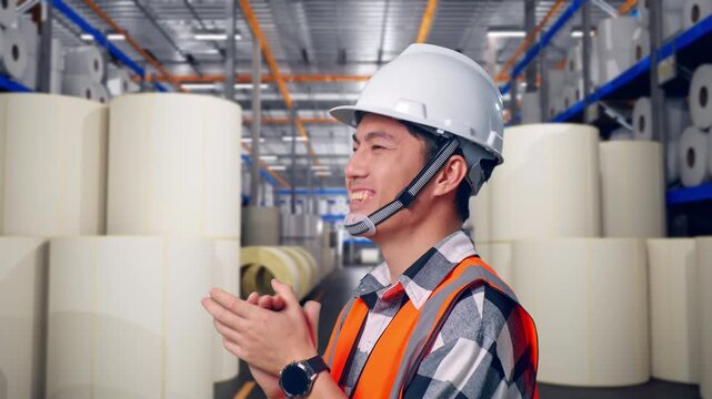 Close Up Side View Of Asian Male Engineer With Safety Helmet Smiling And Clapping His Hands While Standing at Warehouse with Rows of Stacked White Polymer and Paper Rolls