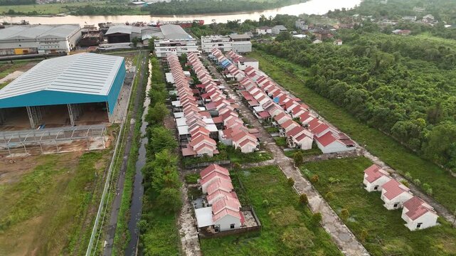 Aerial drone view of a planned housing development by a river, showing orderly rows of red-roofed small houses, adjacent warehouse and green buffer lands.