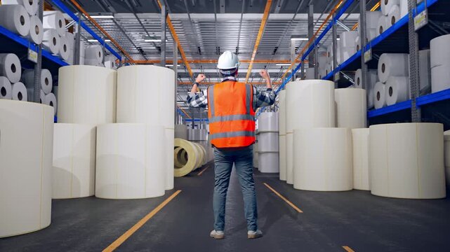 Full Body Back View Of A Male Engineer With Safety Helmet Raising His Hands Celebrating While Working at Warehouse with Rows of Stacked White Polymer and Paper Rolls