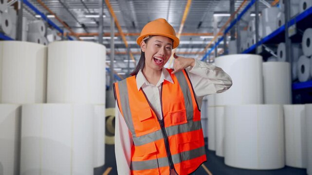 Asian Female Engineer With Safety Helmet Smiling To Camera And Making Call Me Gesture While Standing at Warehouse with Rows of Stacked White Polymer and Paper Rolls