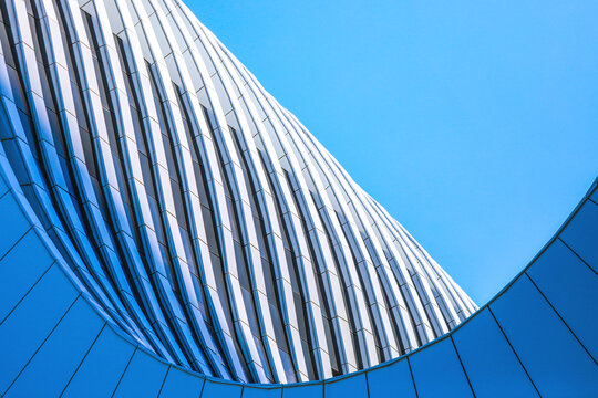 Modern building facade featuring dynamic architectural curves and flowing lines under a bright blue sky, creating an abstract geometric pattern with sleek steel and glass panels
