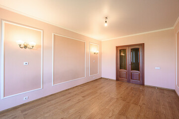 Fototapeta premium spacious, empty room with light pink walls, white decorative molding, wooden laminate floor, wall sconces, double wooden door with glass panels, and a ceiling light. Clean and ready for furnishing