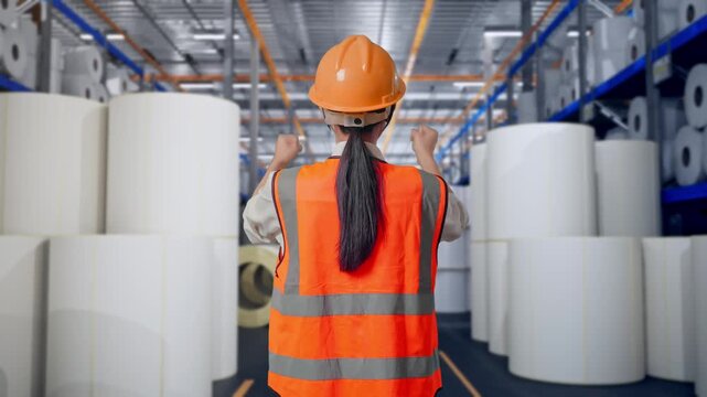 Back View Of A Female Engineer With Safety Helmet Raising Her Hands Celebrating While Working at Warehouse with Rows of Stacked White Polymer and Paper Rolls