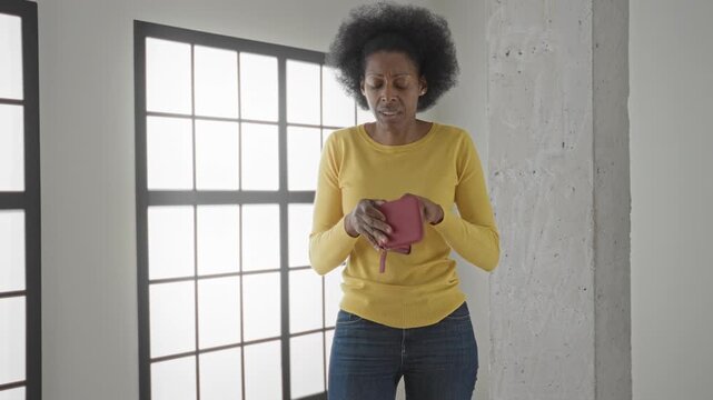 Woman holds empty wallet in hands, shrugs while showing the open wallet inside a building; financial insecurity despair.