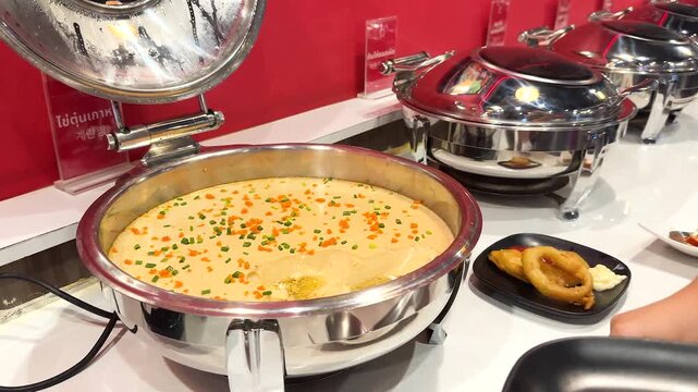 Person serving steamed egg dish from buffet warmer with large metal spoon at restaurant.