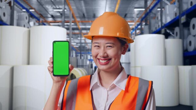 Close Up Of Asian Female Engineer With Safety Helmet Smiling And Showing Green Screen Smartphone To The Camera at Warehouse with Rows of Stacked White Polymer and Paper Rolls