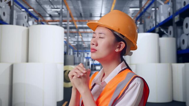 Close Up Side View Of Asian Female Engineer With Safety Helmet Pray For Something While Standing at Warehouse with Rows of Stacked White Polymer and Paper Rolls