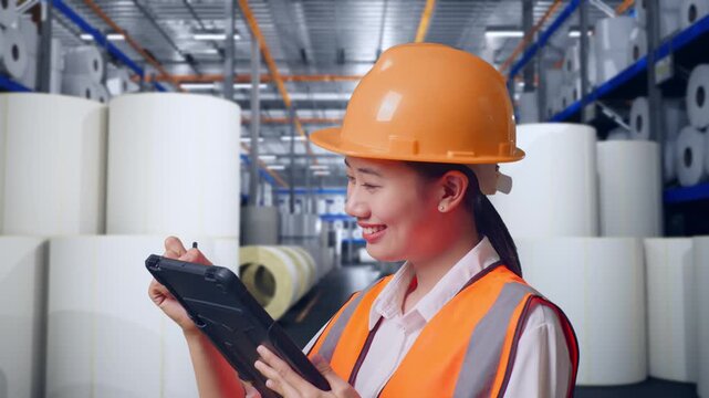 Close Up Side View Of Asian Female Engineer With Safety Helmet Taking Note On The Tablet And Looking Around While Standing at Warehouse with Rows of Stacked White Polymer and Paper Rolls