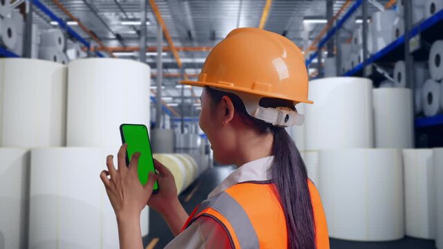 Close Up Back View Of Asian Female Engineer With Safety Helmet Working On A Green Screen Smartphone And Looking Around at Warehouse with Rows of Stacked White Polymer and Paper Rolls