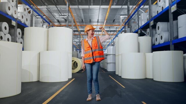 Full Body Of Asian Female Engineer With Safety Helmet Flexing Her Bicep And Smiling To Camera While Standing at Warehouse with Rows of Stacked White Polymer and Paper Rolls