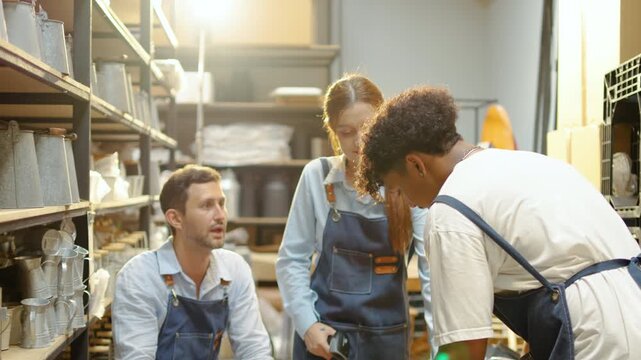 Happy diverse small business team in aprons working in a craft pottery warehouse. Coworkers receive an inventory box, doing a fist bump to celebrate great teamwork and successful store management.