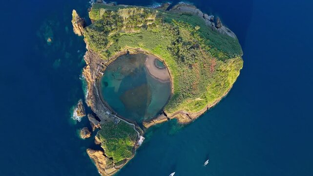 Circular islet lagoon bordered by steep volcanic cliffs rising above Atlantic ocean water in Azores, drone footage