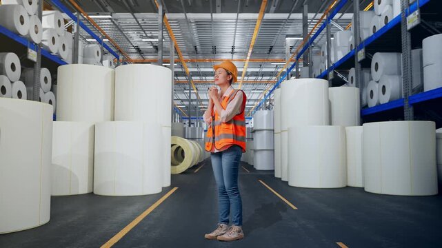 Full Body Side View Of Asian Female Engineer With Safety Helmet Pray For Something While Standing at Warehouse with Rows of Stacked White Polymer and Paper Rolls