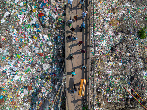 Dhaka, Bangladesh - 11 April 2026: Aerial view of people walking across a narrow bridge over a massive accumulation of plastic waste and garbage on the Buriganga River.