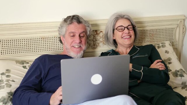 Elderly husband and wife enjoying watching a movie online on a computer together in the bedroom